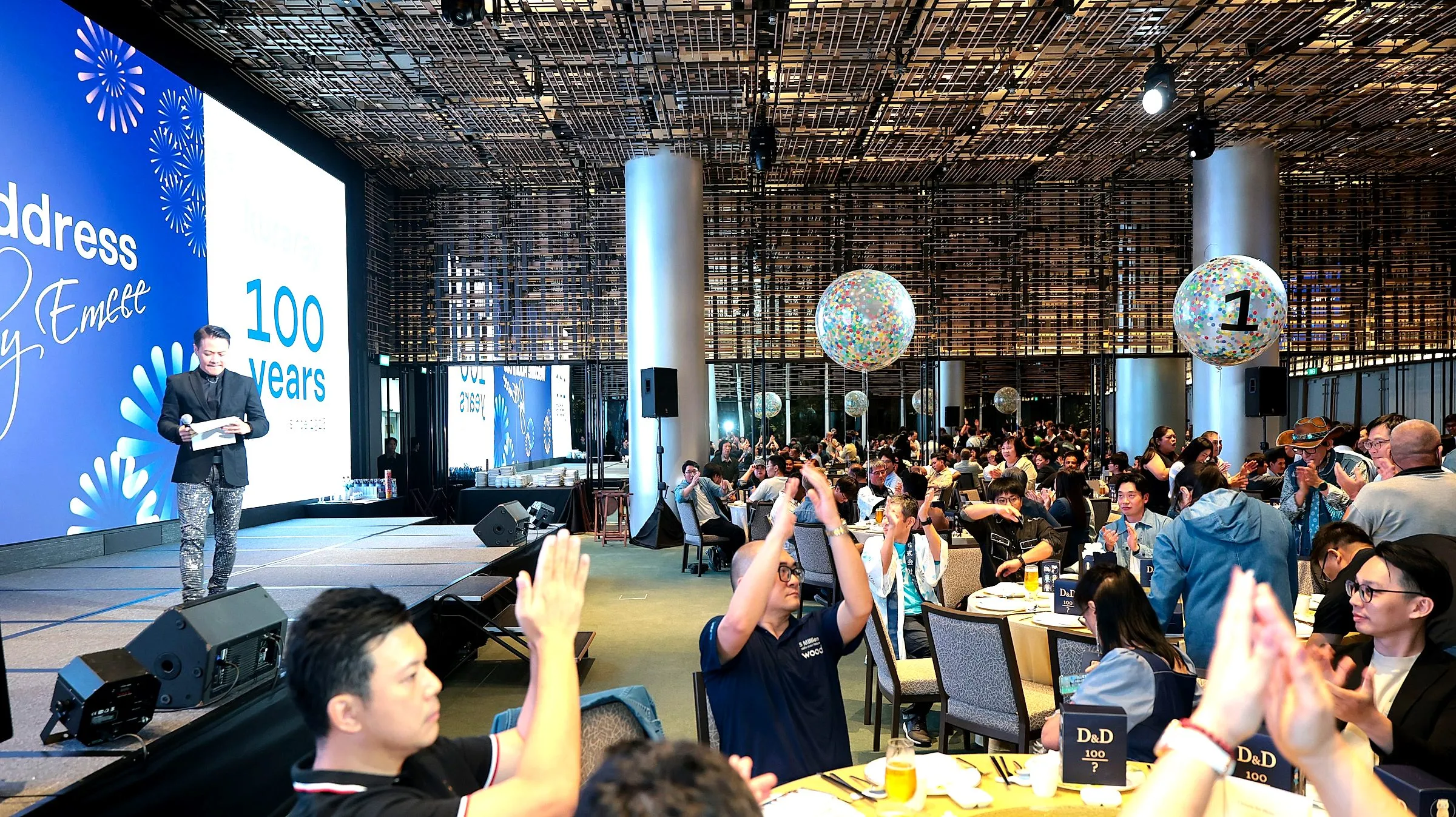 Host on a ballroom stage beside a large screen as guests photograph the moment